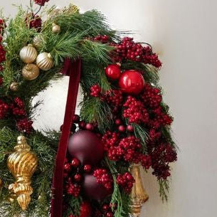 Decorative Christmas wreath with greenery, red berries, gold ornaments, and a red ribbon on a neutral background.